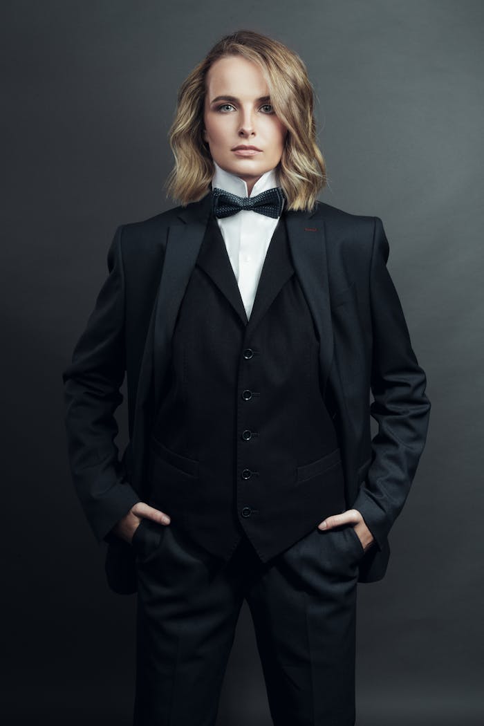 Studio portrait of a person in a tuxedo with a bow tie and hands in pockets, gray background.